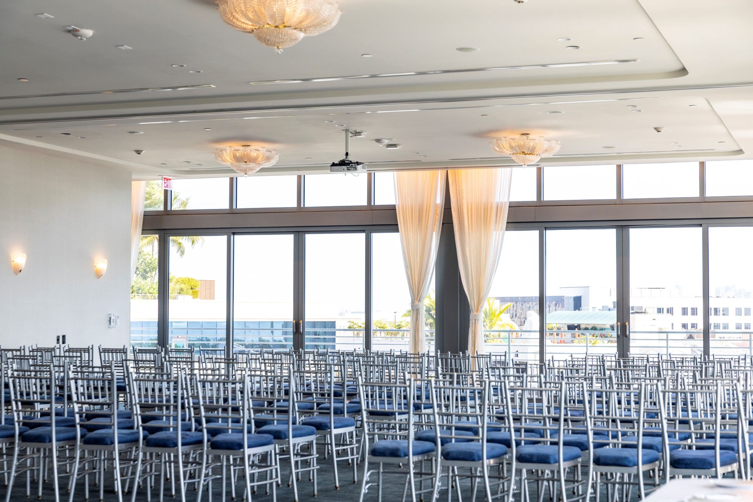 Ampia sala da ballo con file di sedie argentate con cuscini blu rivolte verso grandi vetrate a tutta altezza, Mr. C Miami – Coconut Grove, hotel di lusso.