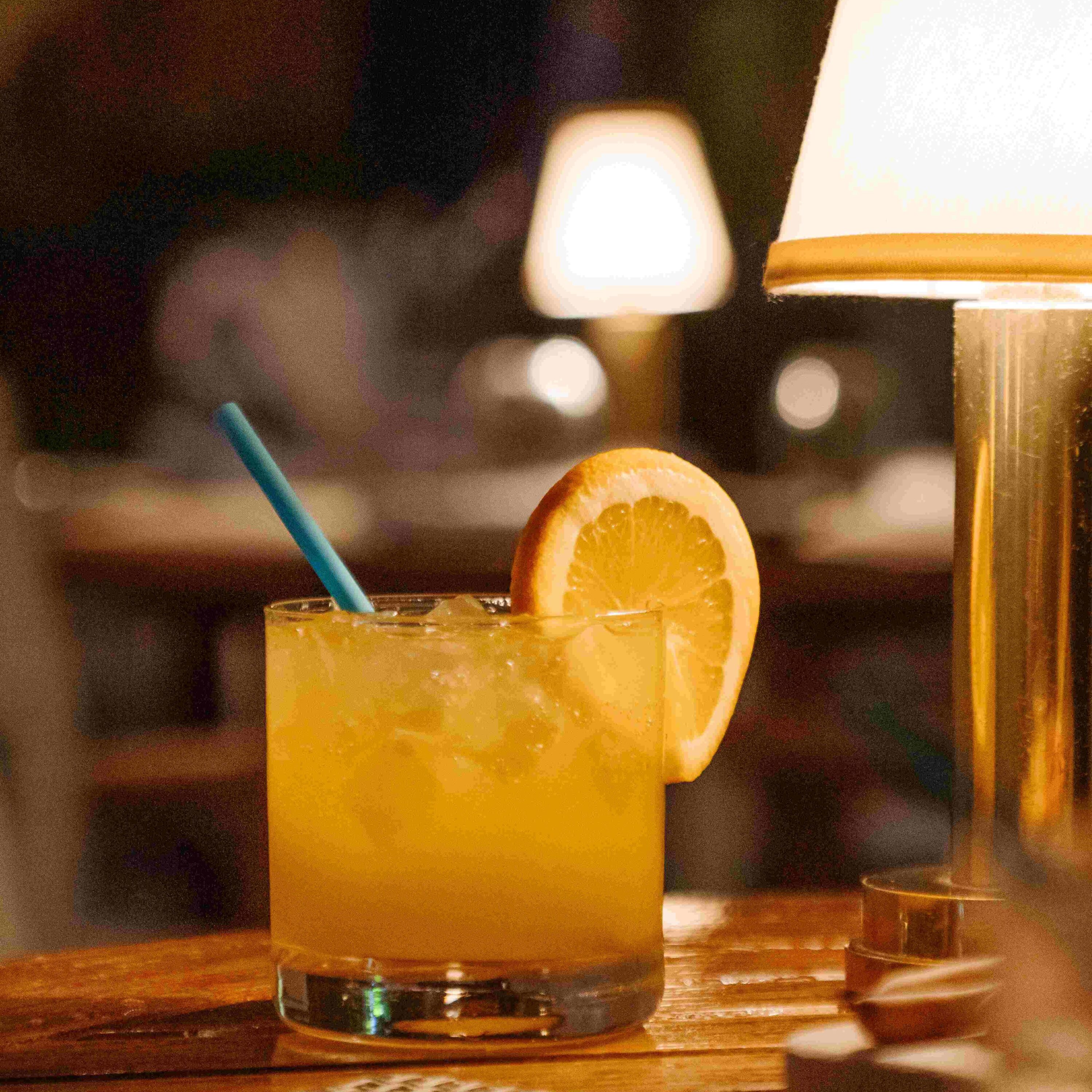 Cocktail jaune orangé avec glaçons et une tranche de citron sur une table en bois à côté d'une lampe chaleureuse, Mr. C Miami – Coconut Grove, hôtel de luxe.