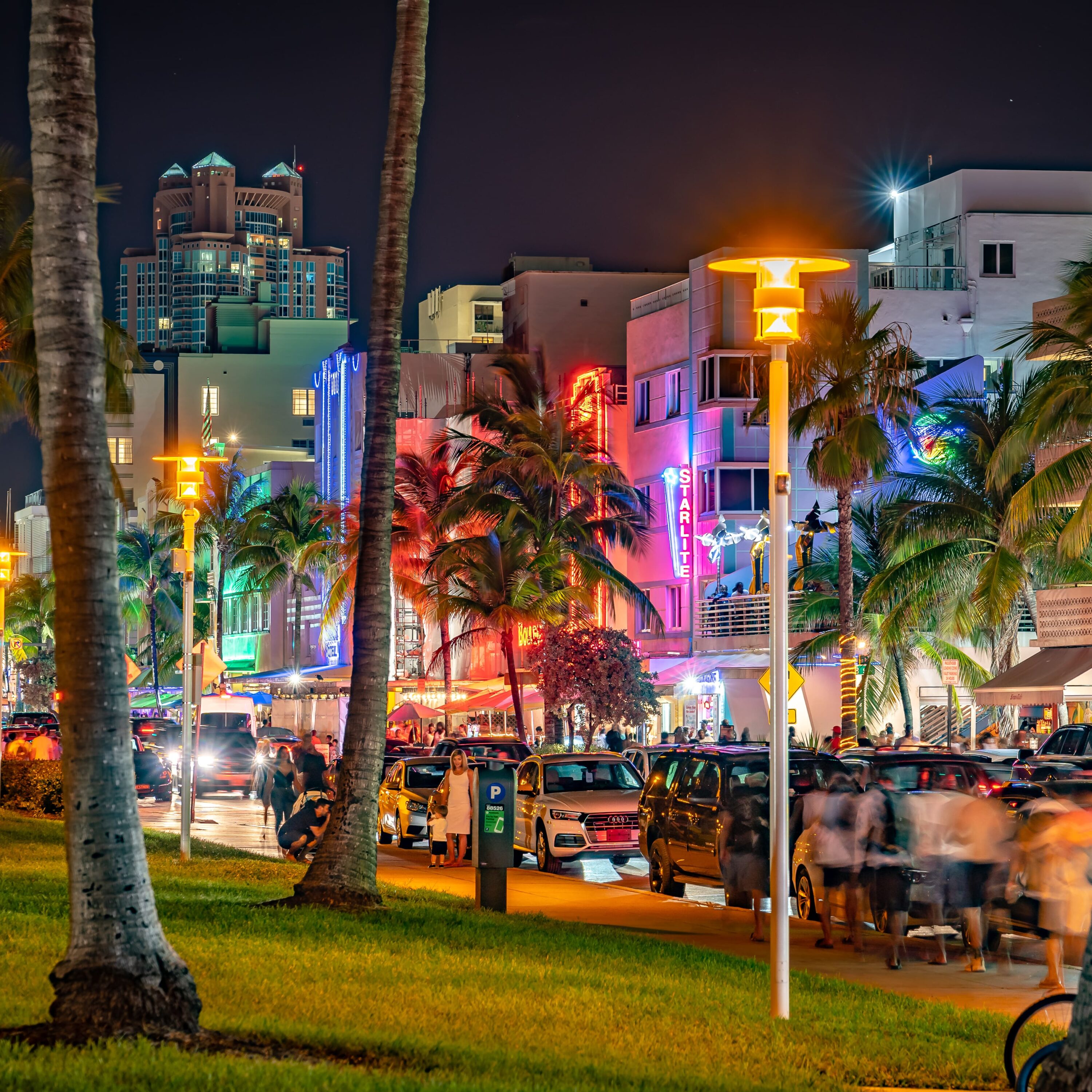 Scène de rue nocturne avec palmiers, bâtiments illuminés au néon, voitures garées et piétons sur le trottoir, Mr. C Miami – Coconut Grove, hôtel de luxe.