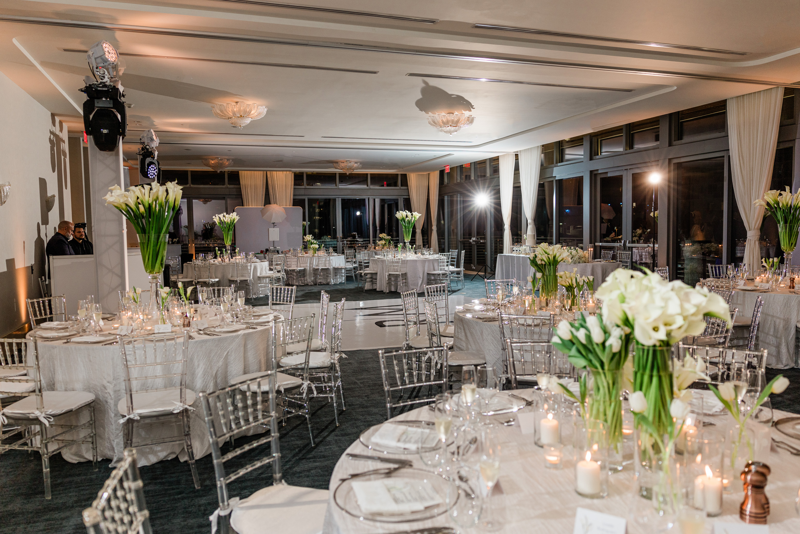 Elegant banquet hall with round tables, clear chairs, white floral centerpieces, and lit candles.