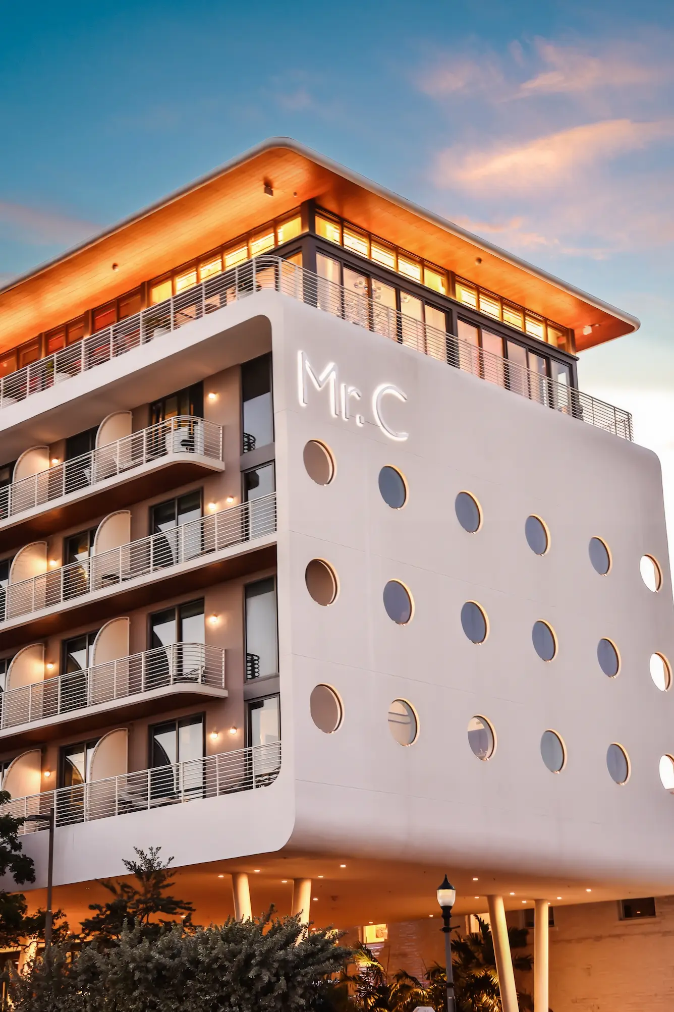 Modern white building with circular windows, balconies, and glowing sign reading Mr. C at sunset , Mr. C Miami – Coconut Grove, luxury hotel.