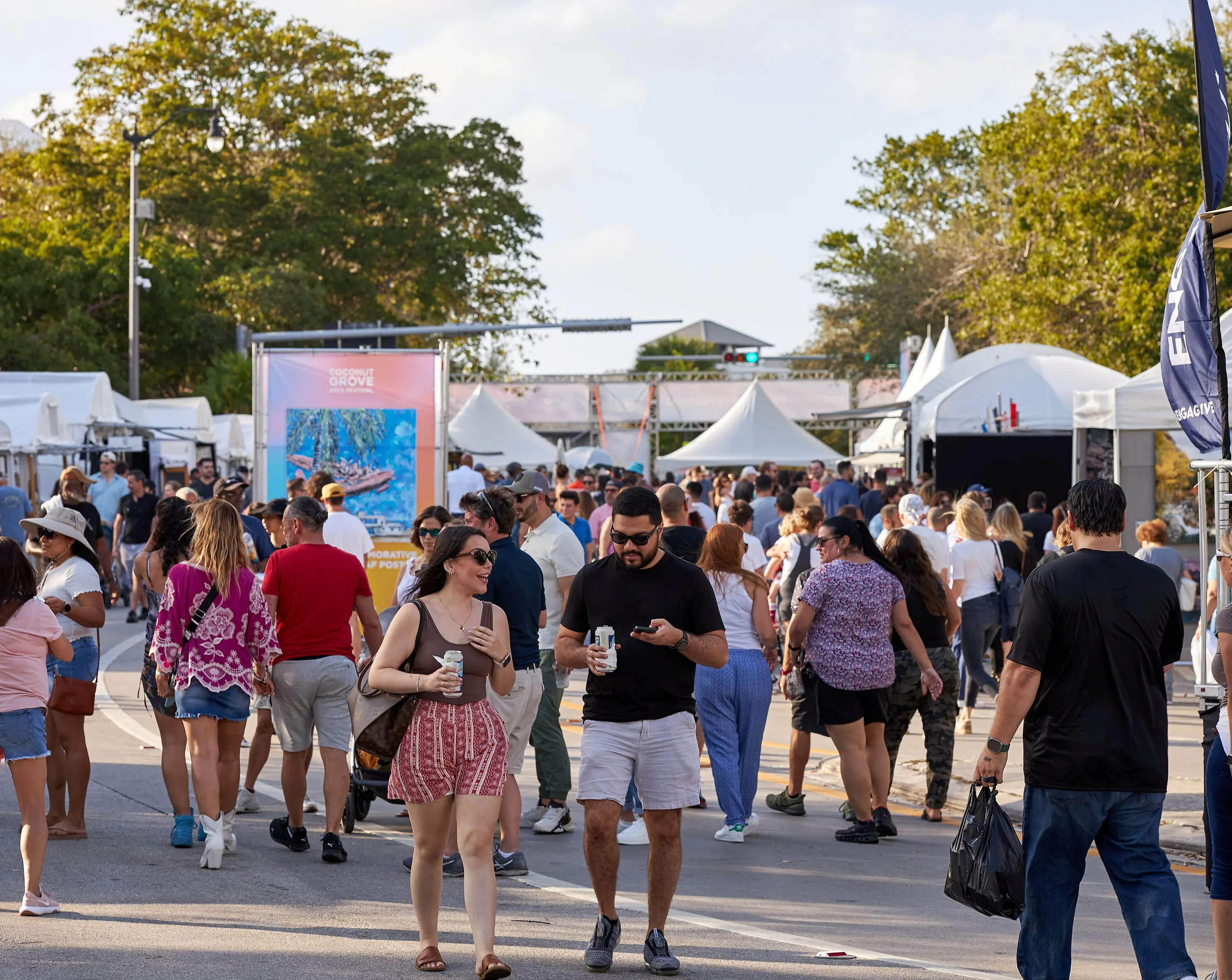Multidão caminhando entre estandes com tendas em um festival de arte ao ar livre sob um céu ensolarado, Mr. C Miami – Coconut Grove, hotel de luxo.