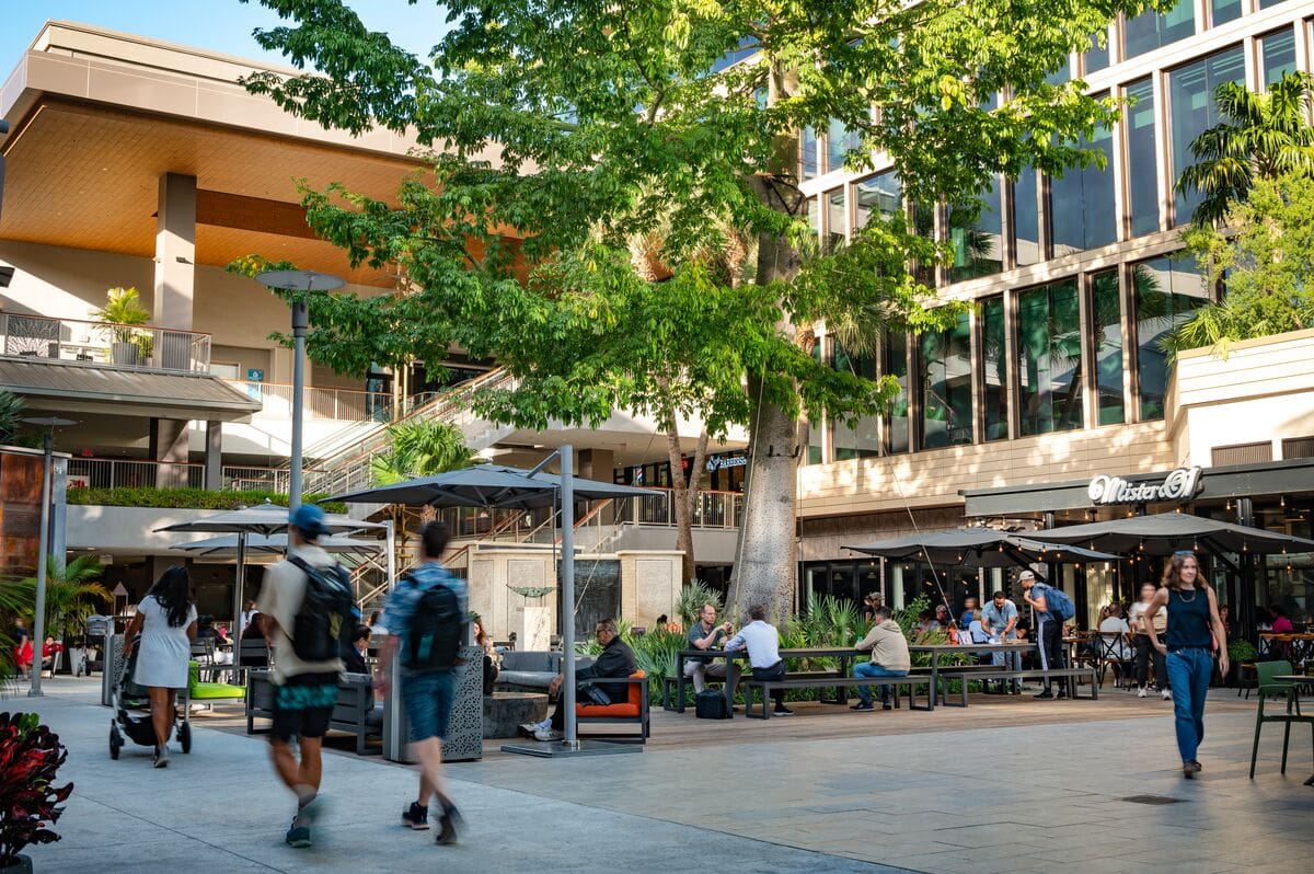 Place urbaine en extérieur avec des personnes qui marchent, assises sous des parasols, et des arbres près d'un bâtiment aux grandes fenêtres, Mr. C Miami – Coconut Grove, hôtel de luxe.