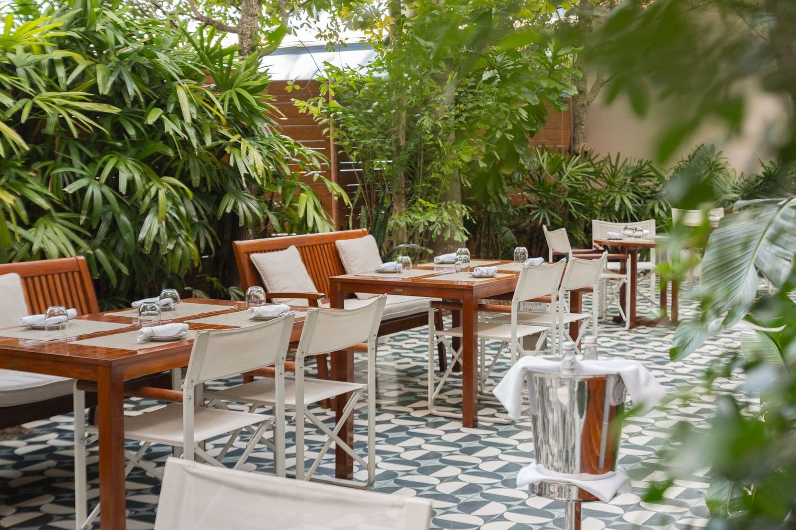 Espace de restauration en extérieur avec tables en bois, chaises blanches et plantes luxuriantes sur un sol en carreaux à motifs, Mr. C Miami – Coconut Grove, hôtel de luxe.