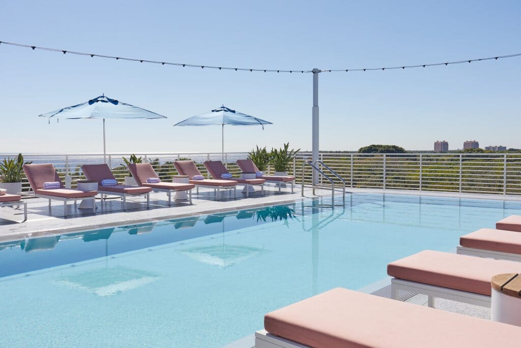 Piscine extérieure avec chaises longues à coussins roses et parasols bleus surplombant la verdure et les bâtiments au loin, Mr. C Miami – Coconut Grove, hôtel de luxe.