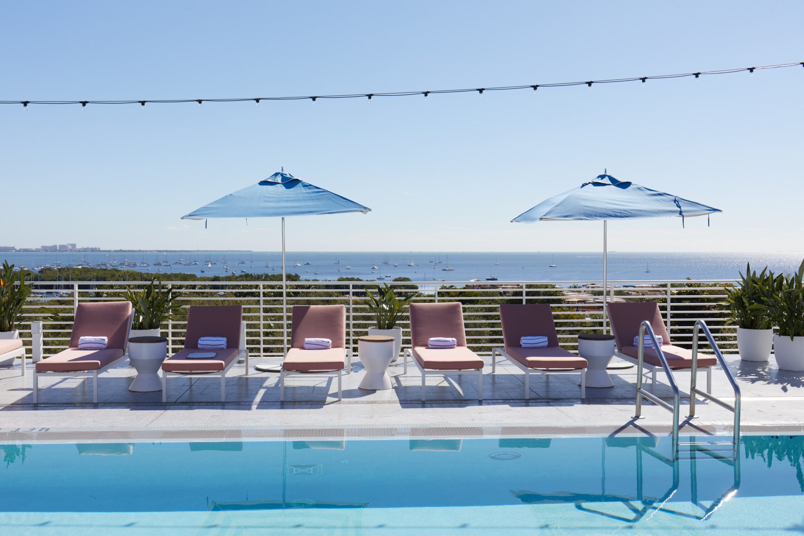 Sedie a sdraio a bordo piscina con asciugamani sotto ombrelloni blu che si affacciano su un oceano calmo con barche, Mr. C Miami – Coconut Grove, hotel di lusso.