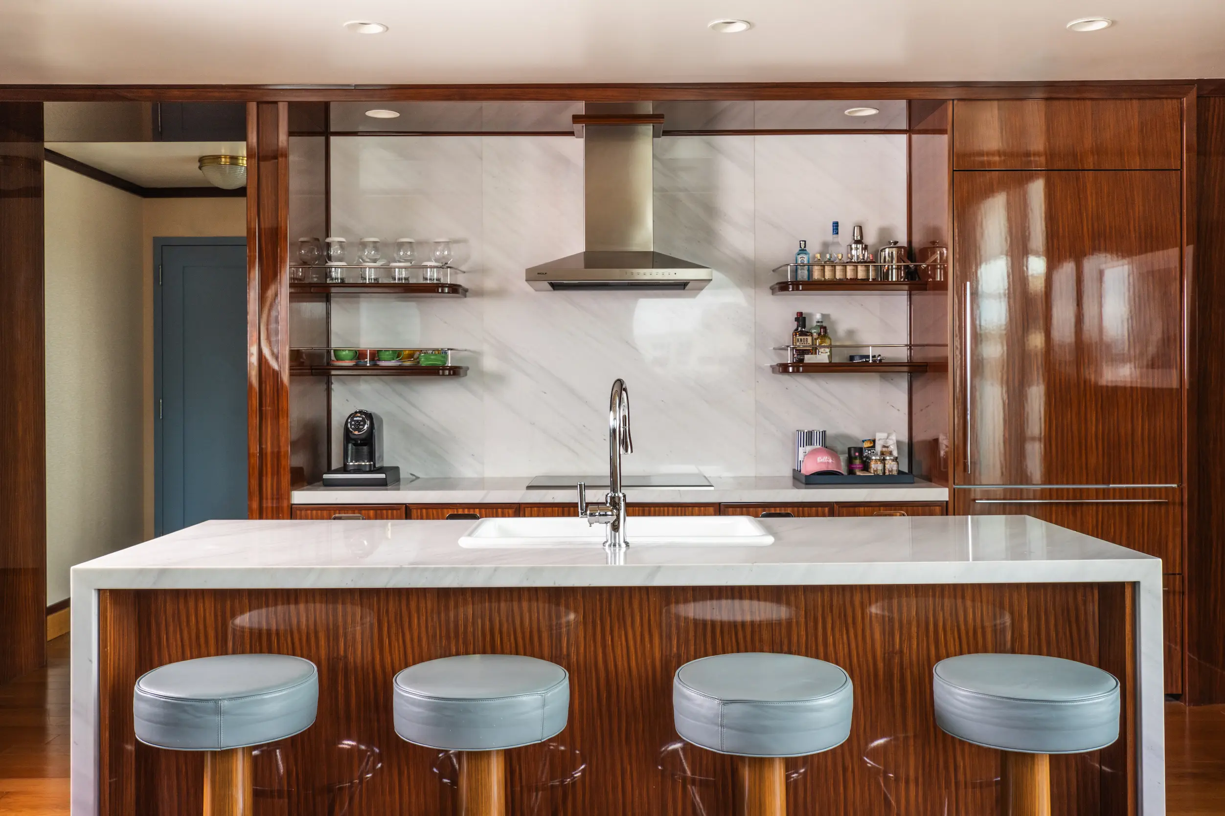 Ilha de cozinha com quatro banquetas azul-claras, voltada para um exaustor de aço inoxidável e prateleiras abertas no Mr. C Miami – Coconut Grove, um hotel de luxo em Miami.