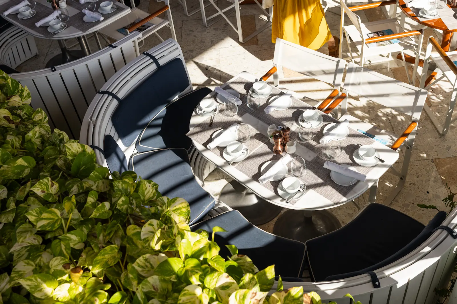 Sunlit restaurant table set with cups, glasses, and napkins, surrounded by curved bench with cushions.