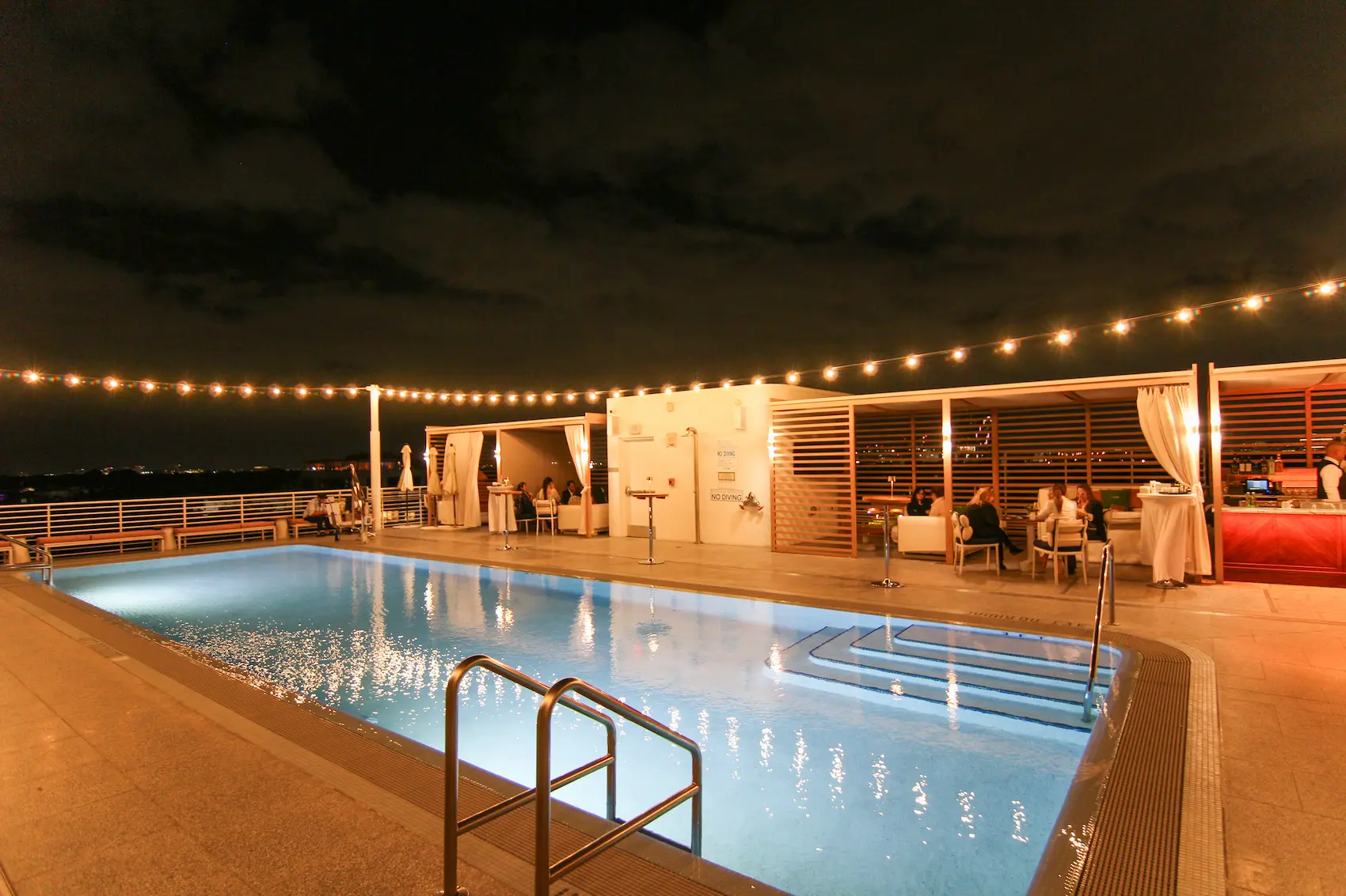 Night view of a lit outdoor pool with string lights and people sitting in cabanas and at tables nearby.