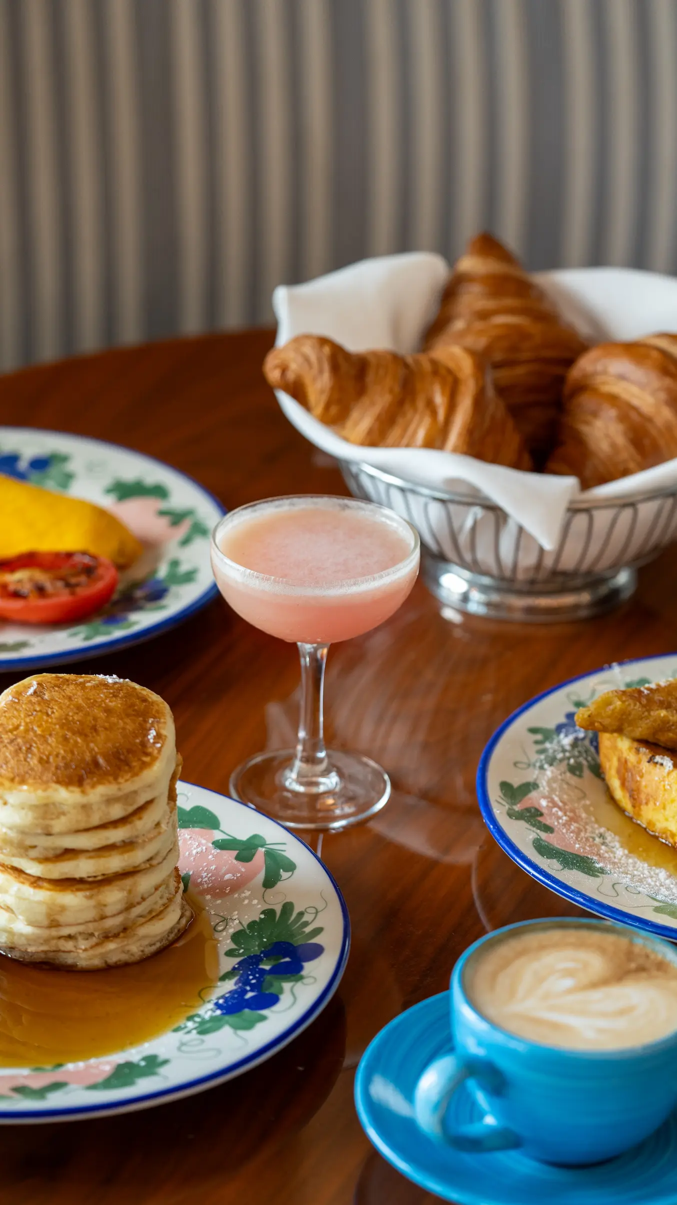 Breakfast table with pancakes, croissants, pink cocktail, coffee, and plates with food on a wooden surface , Mr. C Miami – Coconut Grove, luxury hotel.