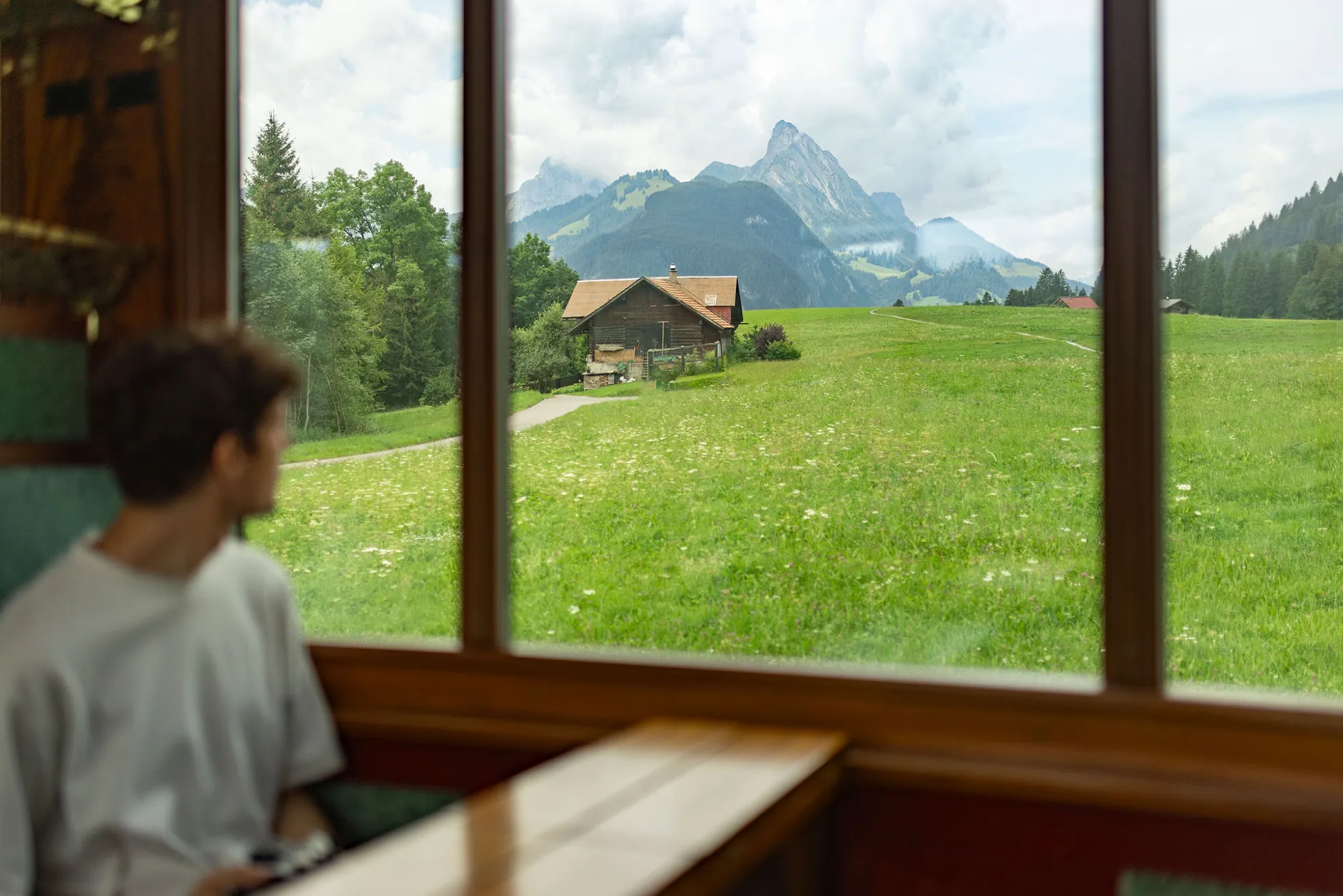 Person inside looking out a large window at green fields, a wooden house, and mountains under a cloudy sky.