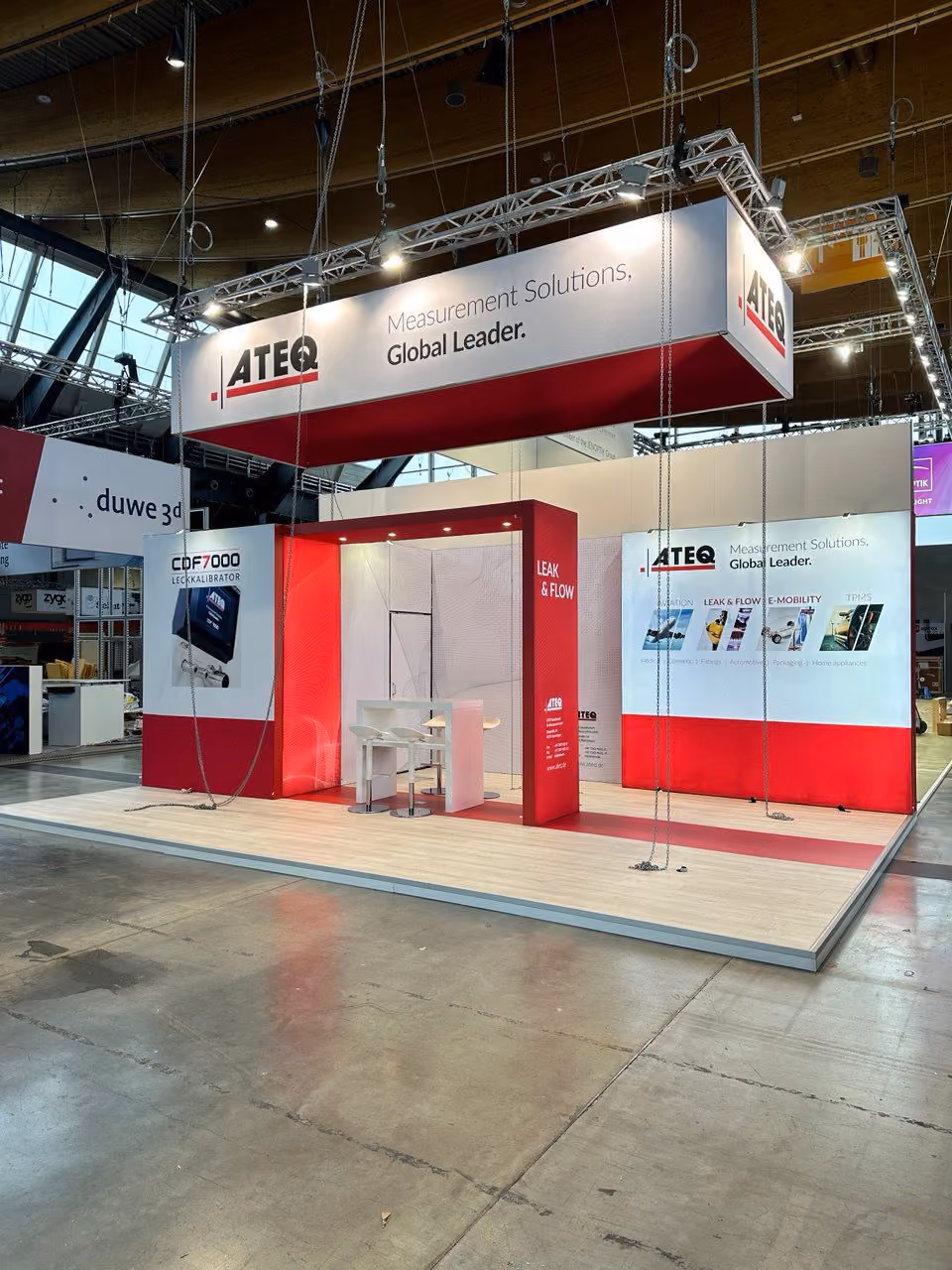ATEQ exhibition stand with red and white branding, overhead hanging signage, product display panels, and meeting area