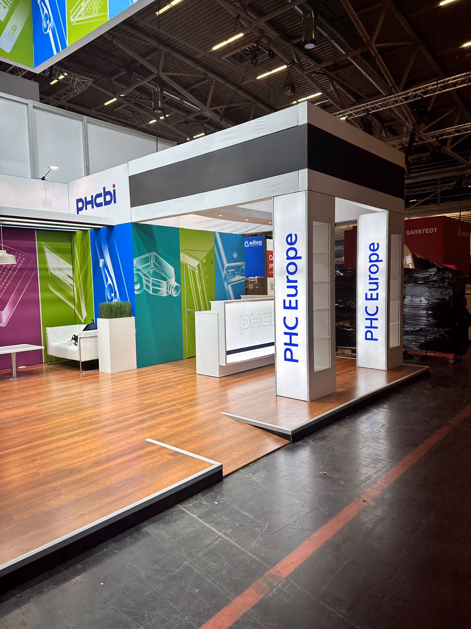 PHC Europe exhibition stand with illuminated overhead branding, colorful graphics, and open product display areas