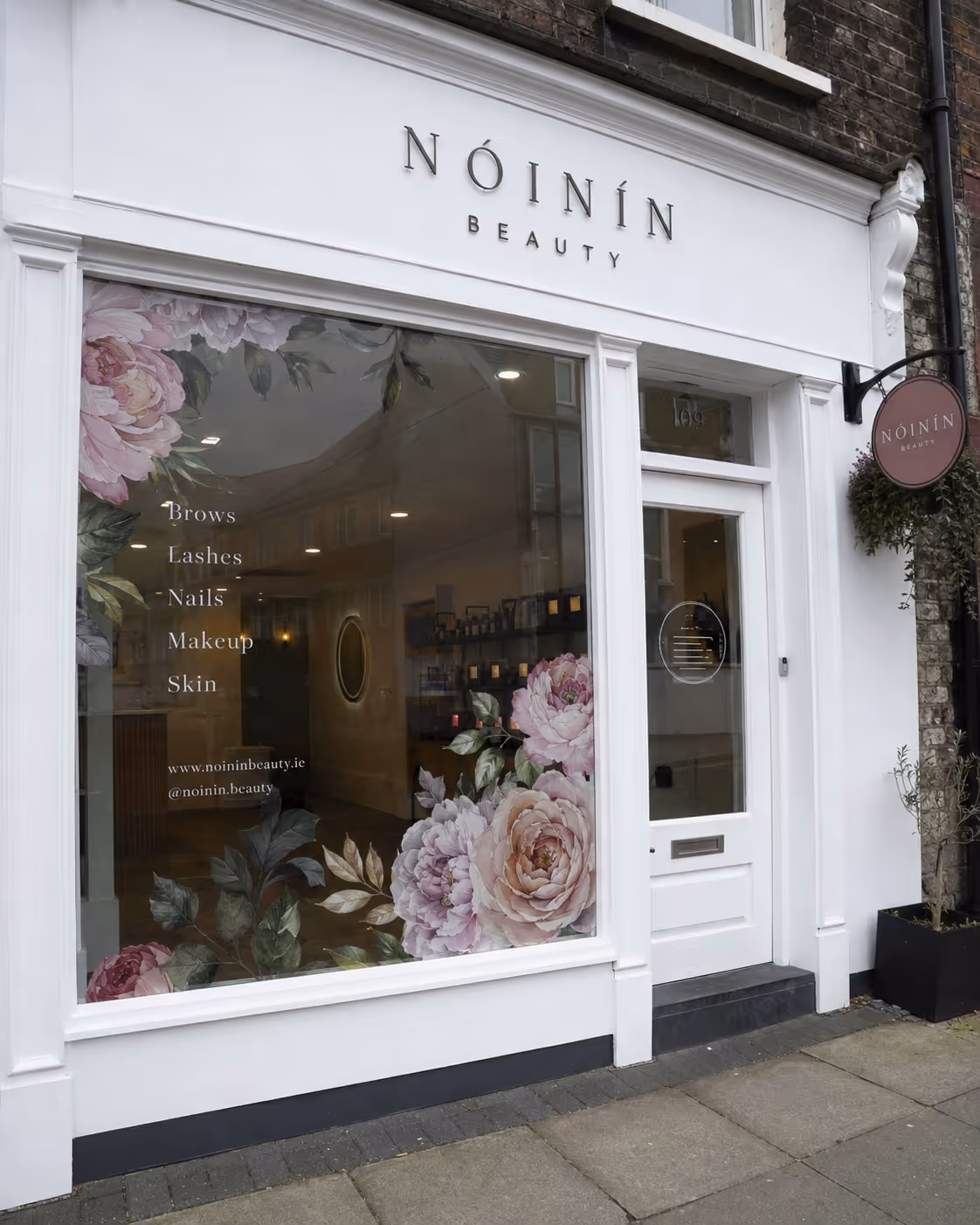 White storefront of Nóinín Beauty with floral window art and services listed: Brows, Lashes, Nails, Makeup, Skin.