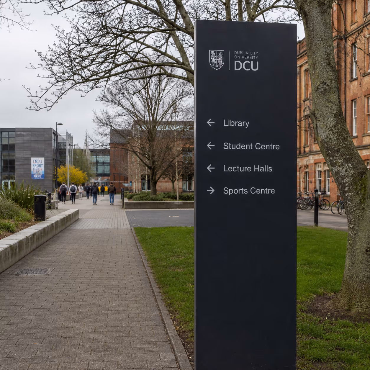 Dark signpost on Dublin City University campus with directions to library, student centre, lecture halls, and sports centre.