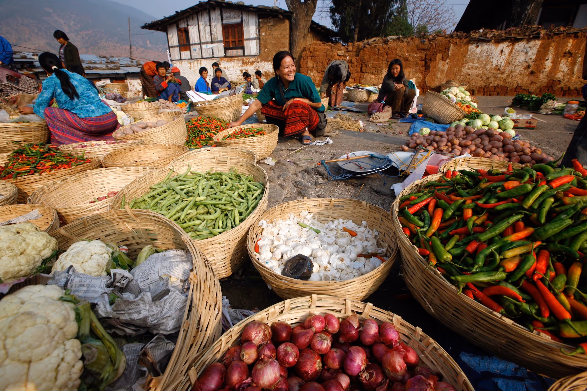 Outdoor market scene with women selling baskets of vegetables including chili peppers, onions, garlic, and cauliflower.