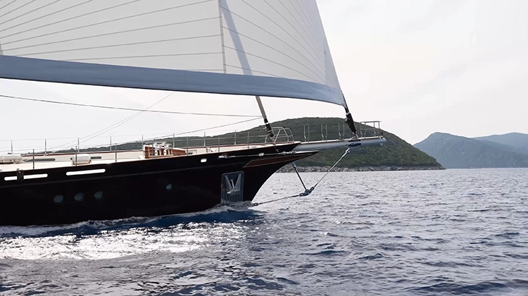Close-up of a sailing yacht bow cutting through calm blue sea near green hilly coastline.
