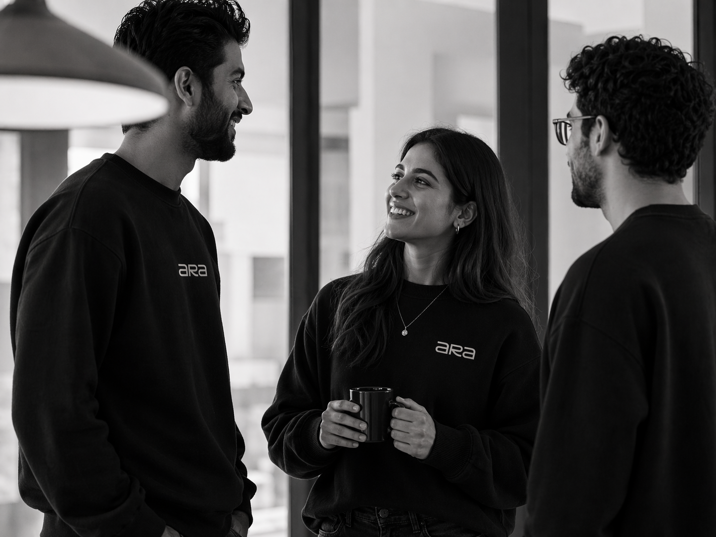 Three people wearing matching Ara sweatshirts talking and smiling indoors, with one woman holding a mug.