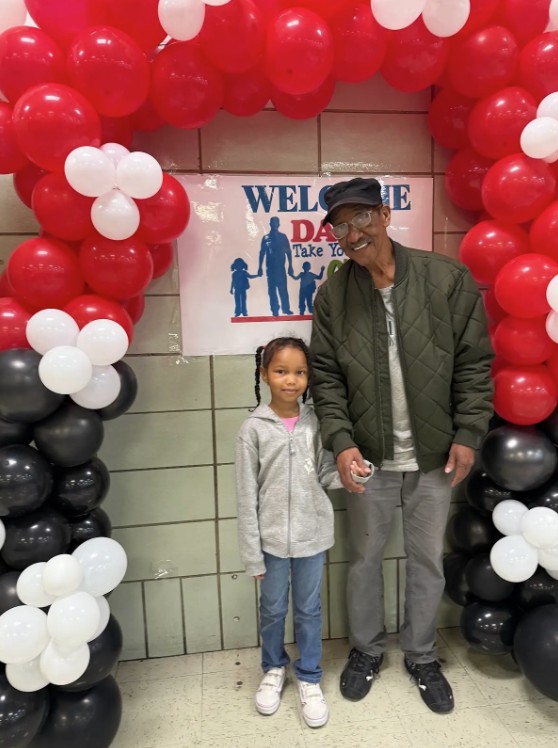 Student and father posing under balloon arch at Dads Take Your Child to School Day