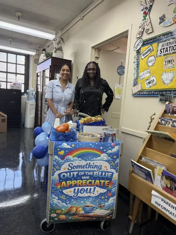 Two staff members smile beside a staff appreciation cart in school hallway