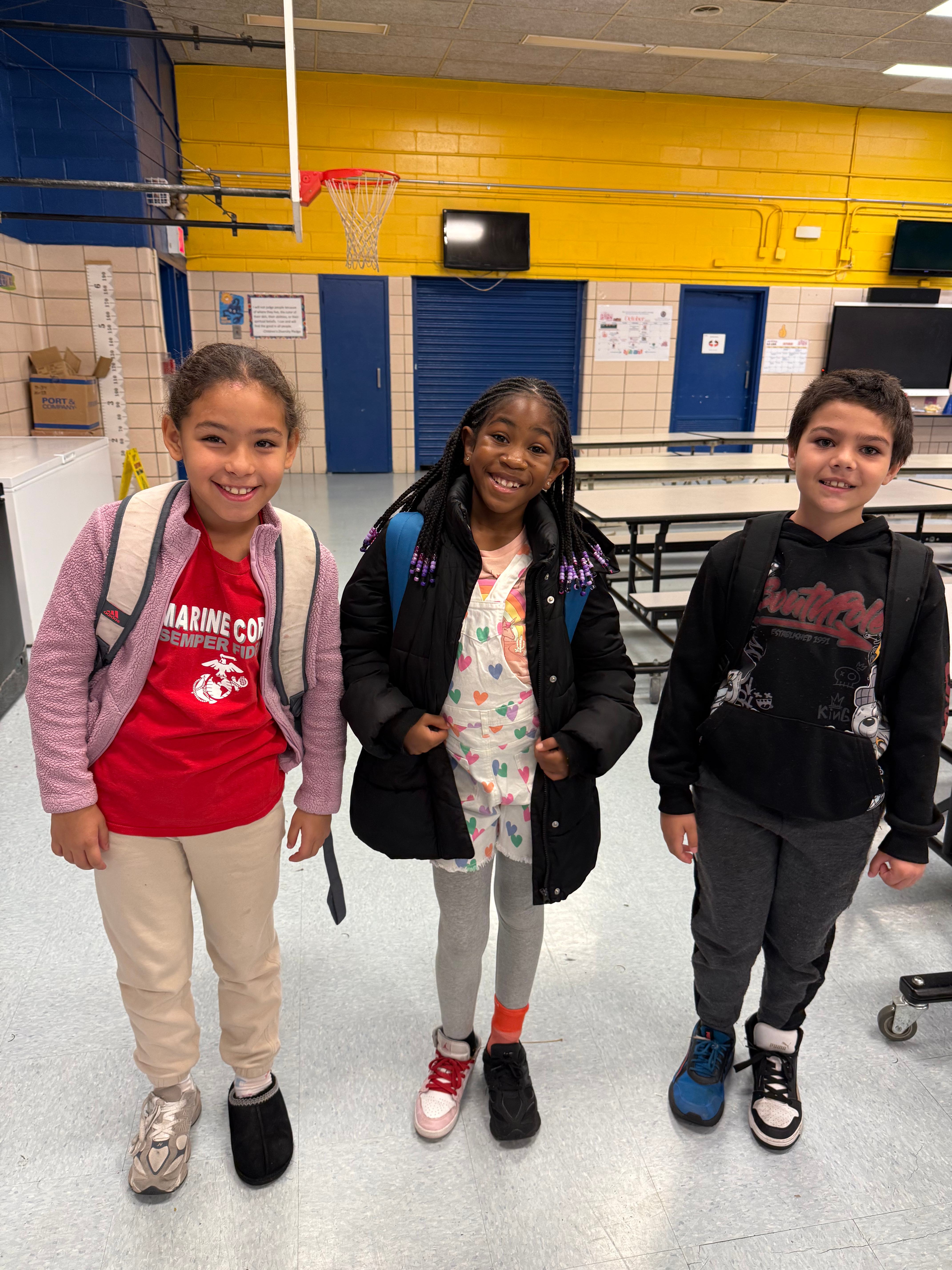 Three smiling elementary students with backpacks in school cafeteria
