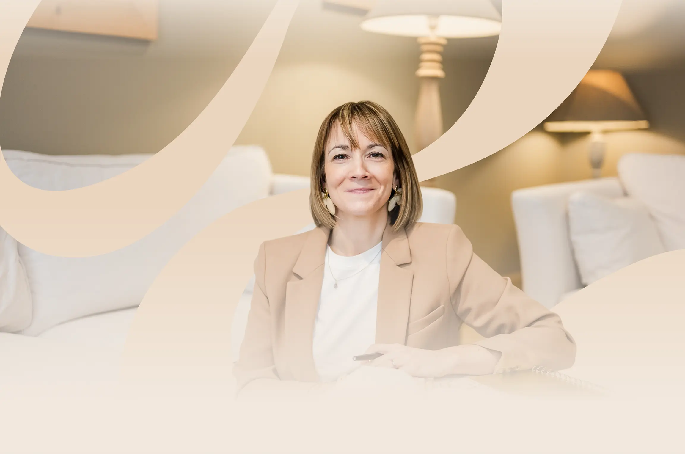 Aurélie Rey assise, portant un haut blanc et une veste beige, le bras gauche posé, sur fond clair avec canapé et coussin en fond