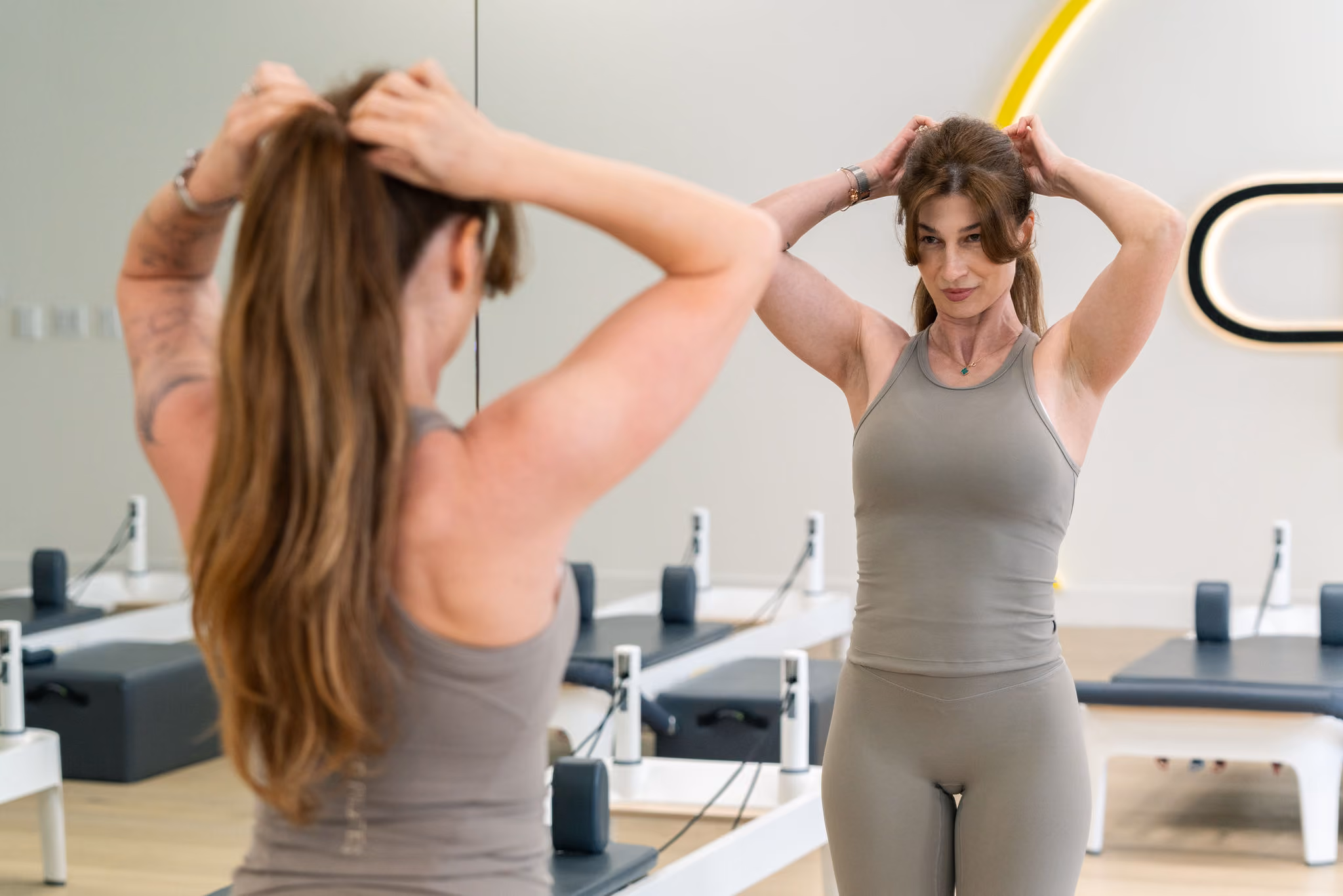 Woman in gray workout clothes tying hair back in front of a mirror in a Pilates studio.