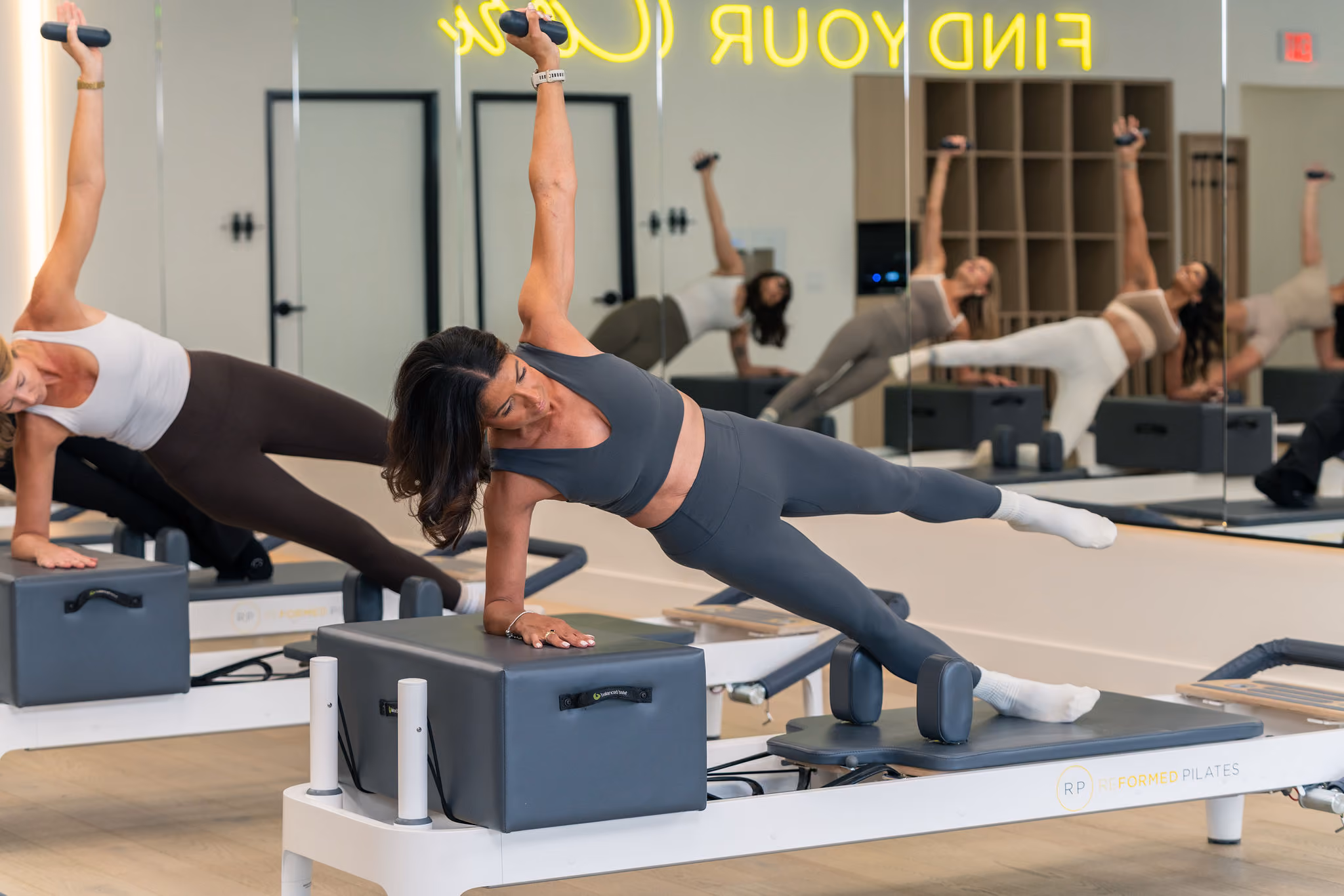 Two women exercising on Pilates reformer machines, balancing on one arm with legs extended.