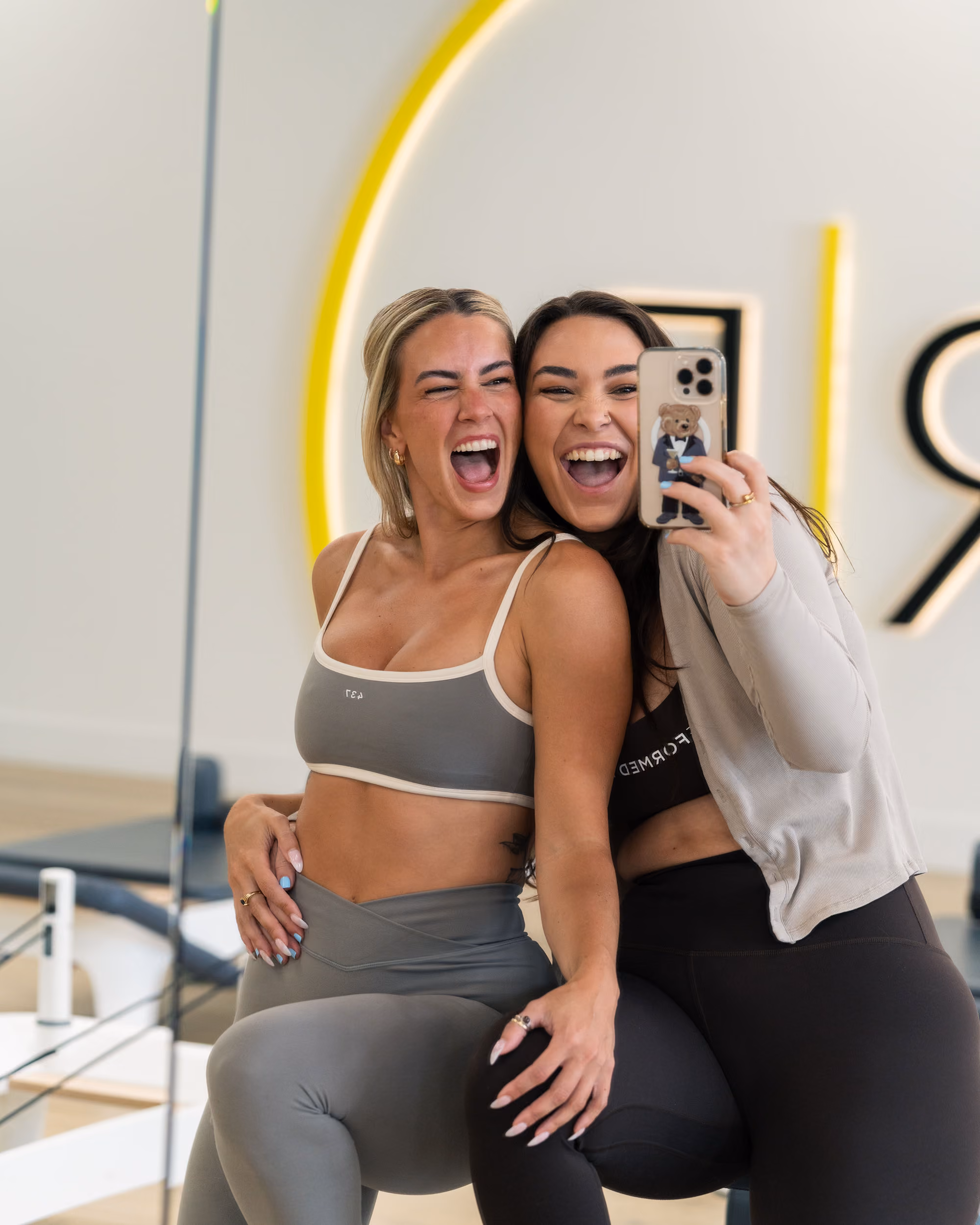 Two women in workout clothes taking a joyful mirror selfie at a fitness studio.