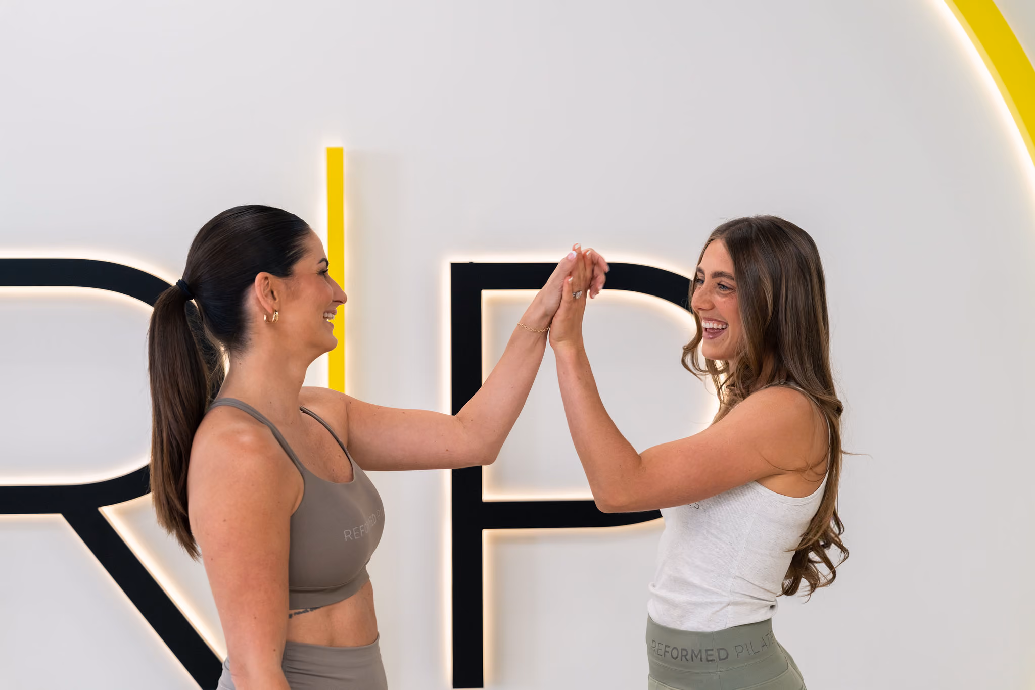 Two women in workout clothes smiling and giving a high five indoors with a Reformed Pilates sign behind.
