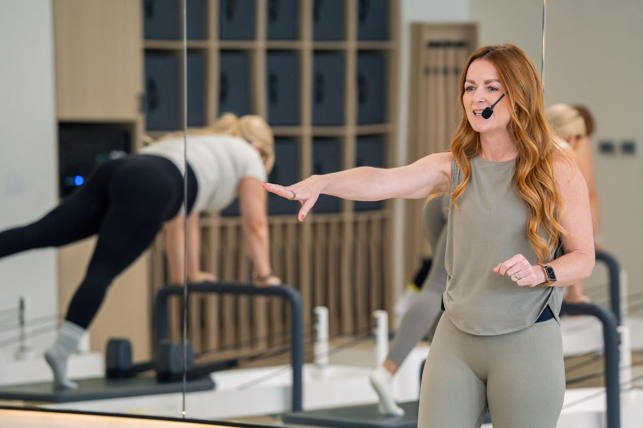 Fitness instructor with headset leads Pilates class while a participant balances on a reformer machine.