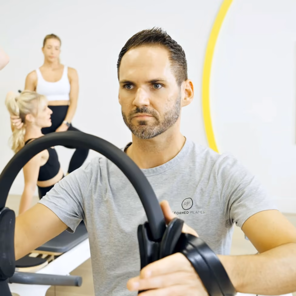Man in gray t-shirt focused on using a Pilates reformer machine in a bright studio with two women in background.