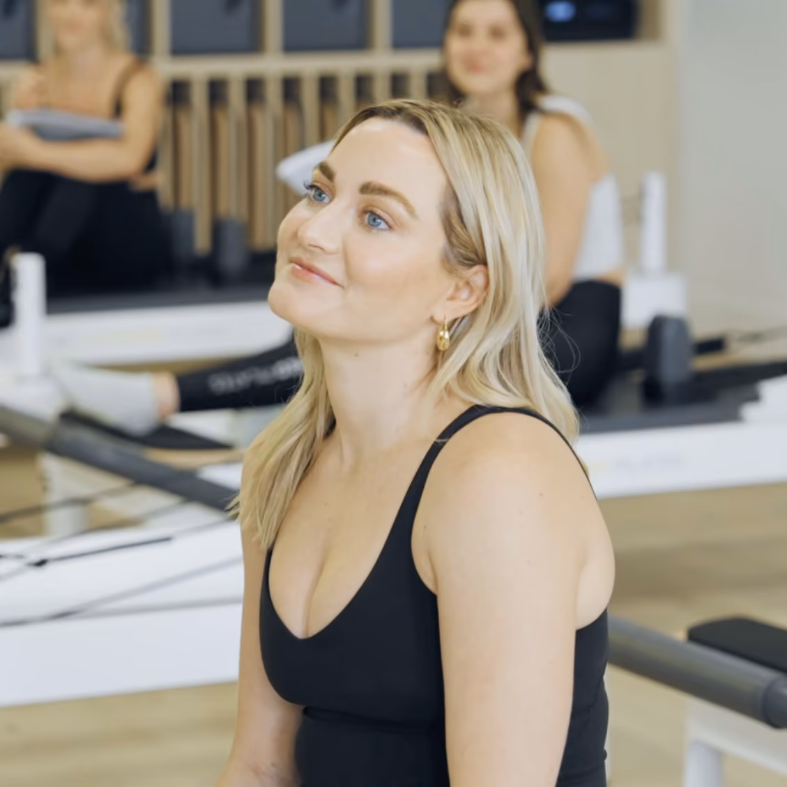 Blonde woman in black tank top sitting and smiling in a gym with two blurred women behind her.