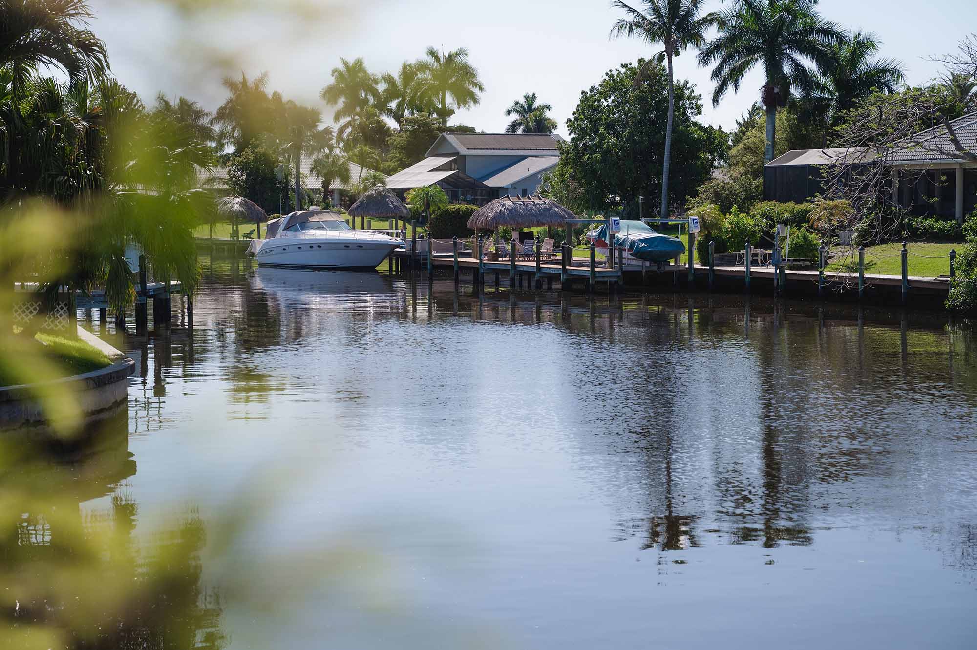 A calm waterfront residential area with boats docked by wooden piers and palm trees along the shore.