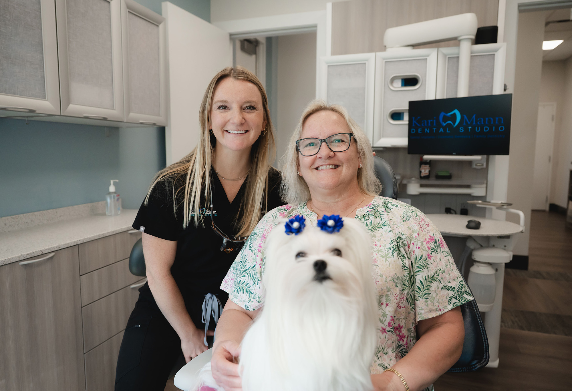 Smiling female dentist and patient seated in dental office with a white dog adorned with blue bows.