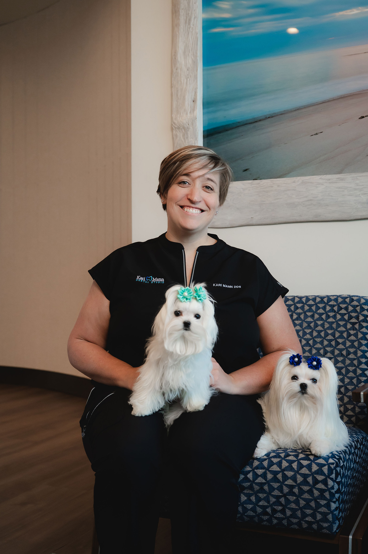 Smiling woman in black uniform sitting with two small white dogs wearing blue bows, one on her lap and one beside her.