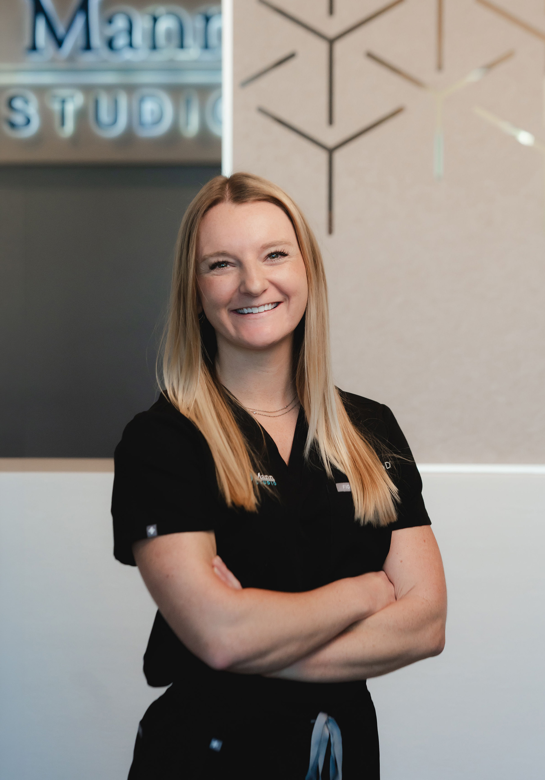 Smiling woman with long blonde hair in black medical scrubs standing with arms crossed indoors.