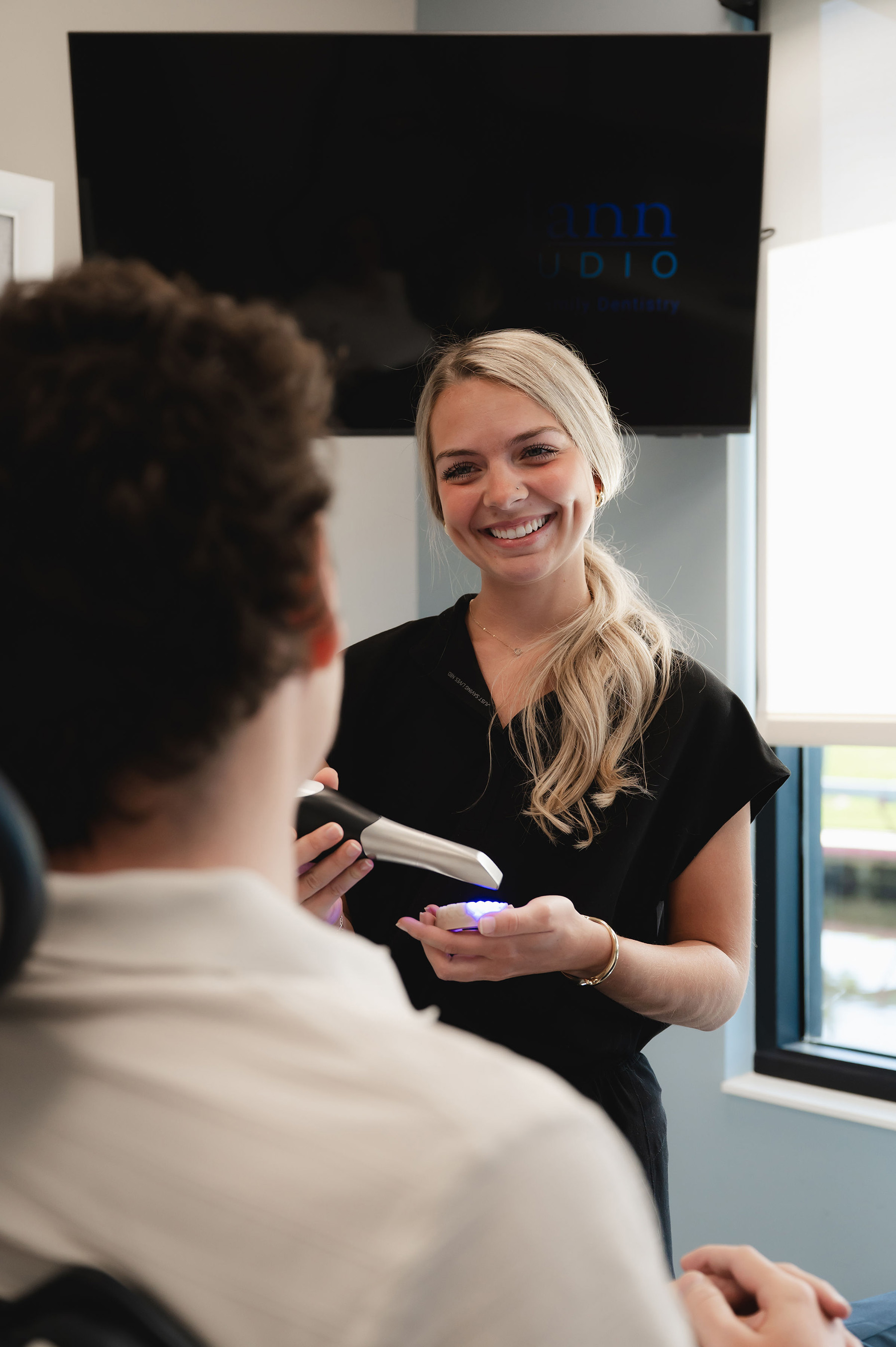 Smiling female dentist holding a dental curing light over a dental mold while talking to a seated patient.