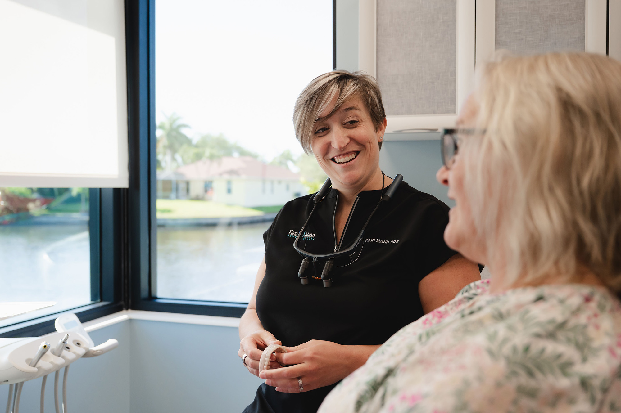 Smiling female dentist holding a dental curing light over a dental mold while talking to a seated patient.