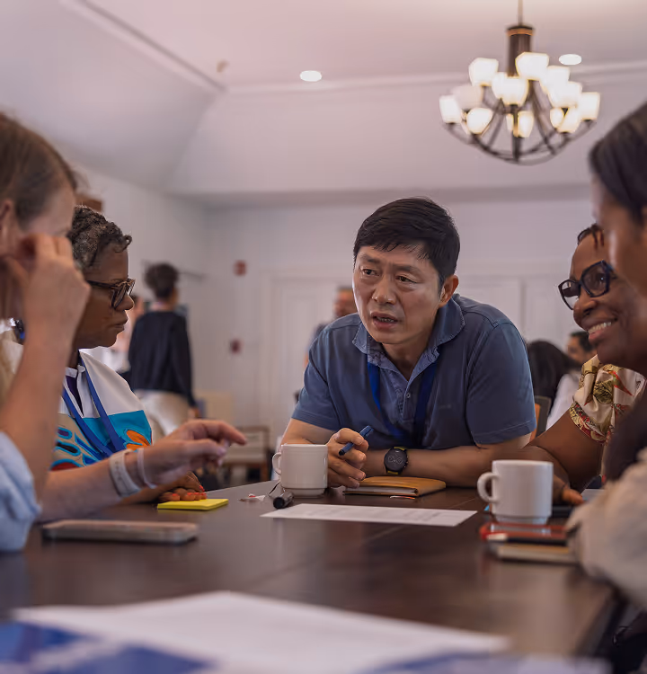 Diverse group of people engaged in a serious discussion around a table with notebooks and mugs.