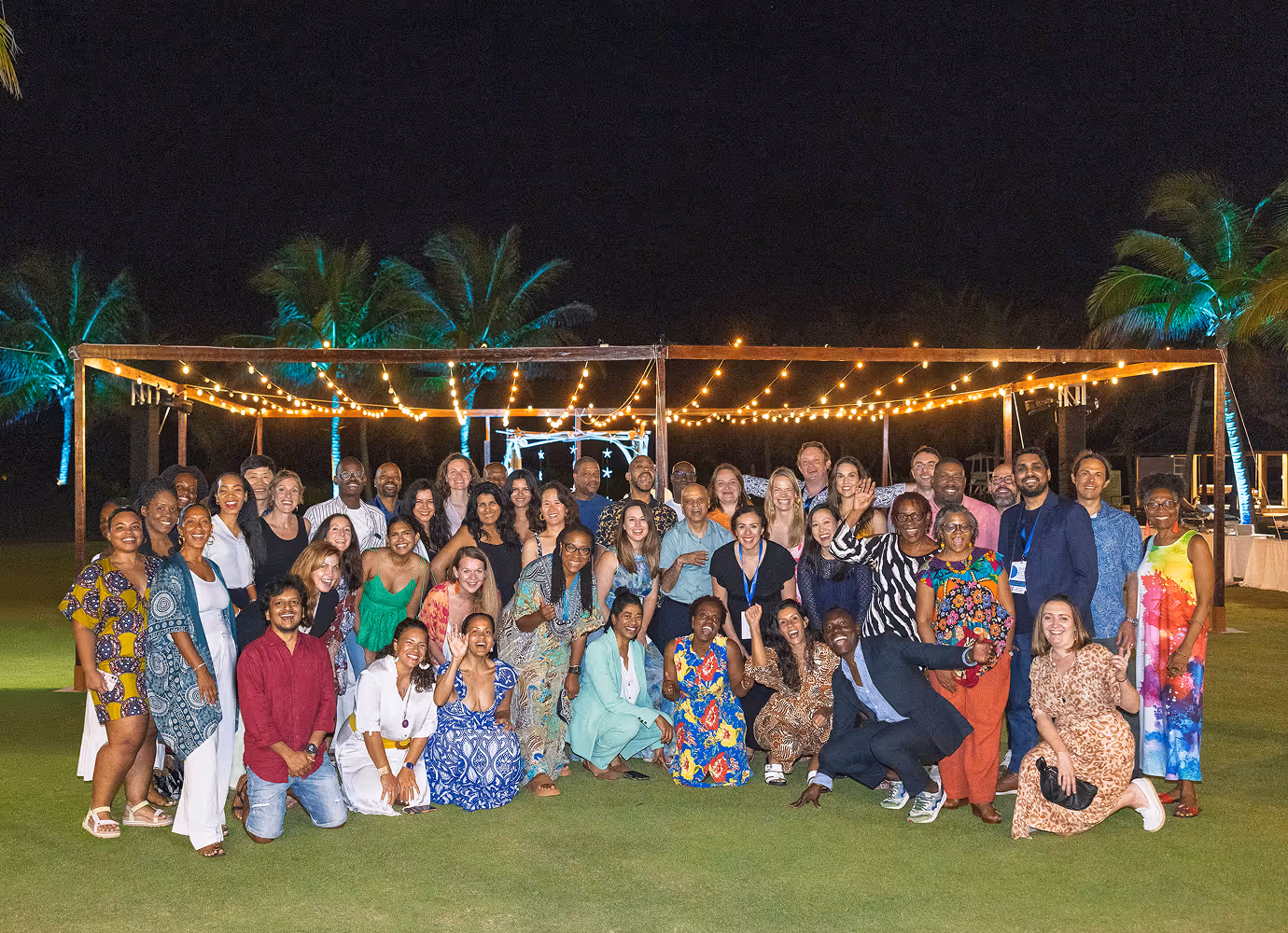 Large diverse group of people smiling and posing outdoors on grass under string lights at night, with palm trees lit in blue in the background.