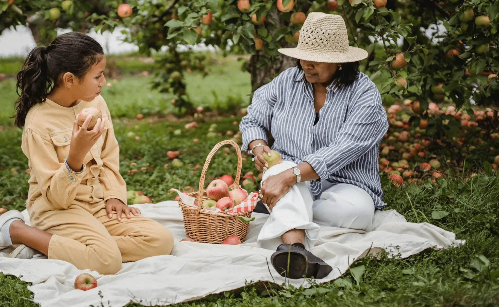 A woman and a girl sitting on a blanket in an apple orchard, sharing apples. The woman wears a hat and striped shirt, while the girl wears a beige outfit. They have a basket full of apples between them.