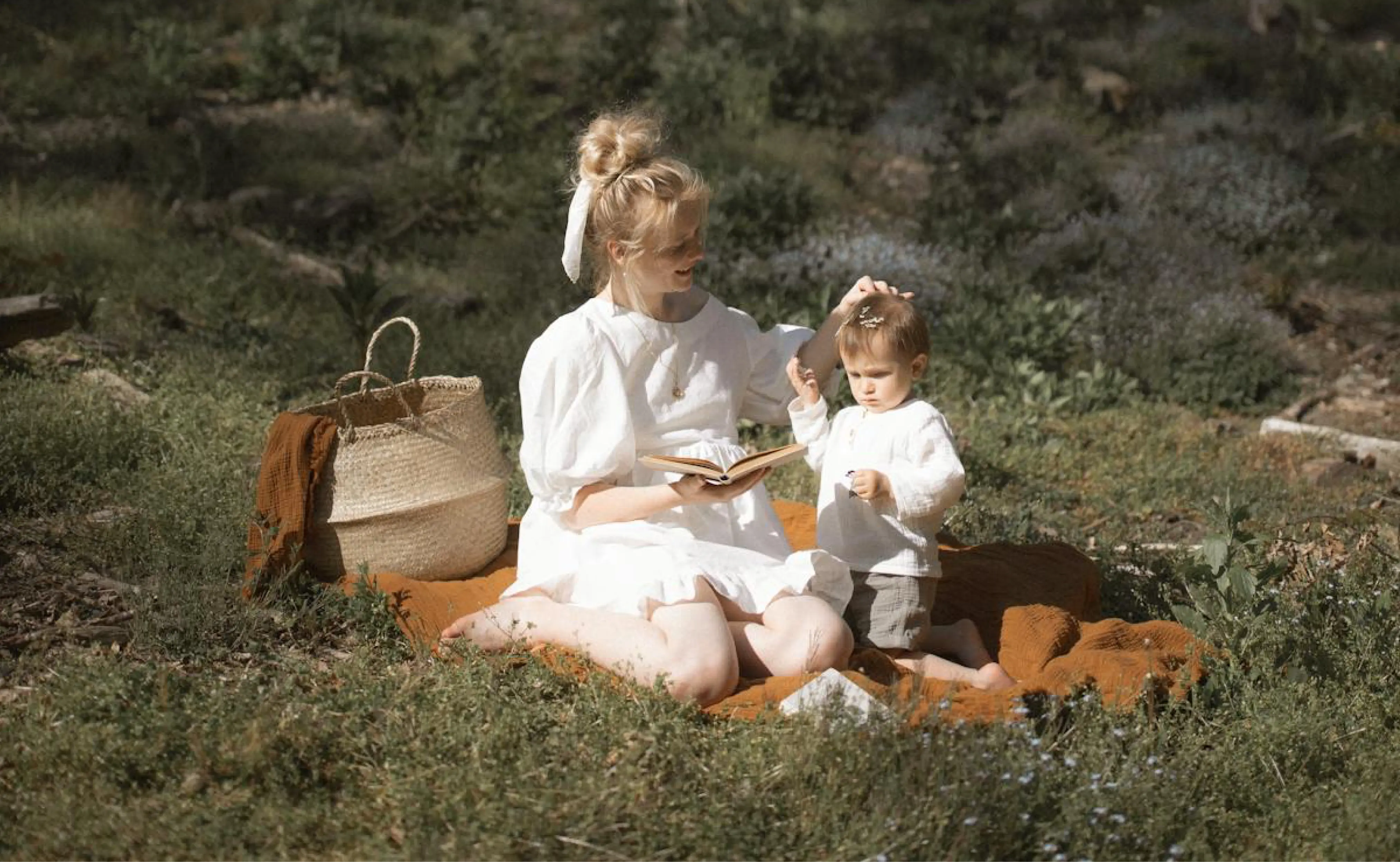 A mother and her young child are sitting on a blanket in a grassy field. The mother is reading a book to the child while gently touching the child's head. They both appear relaxed and happy.
