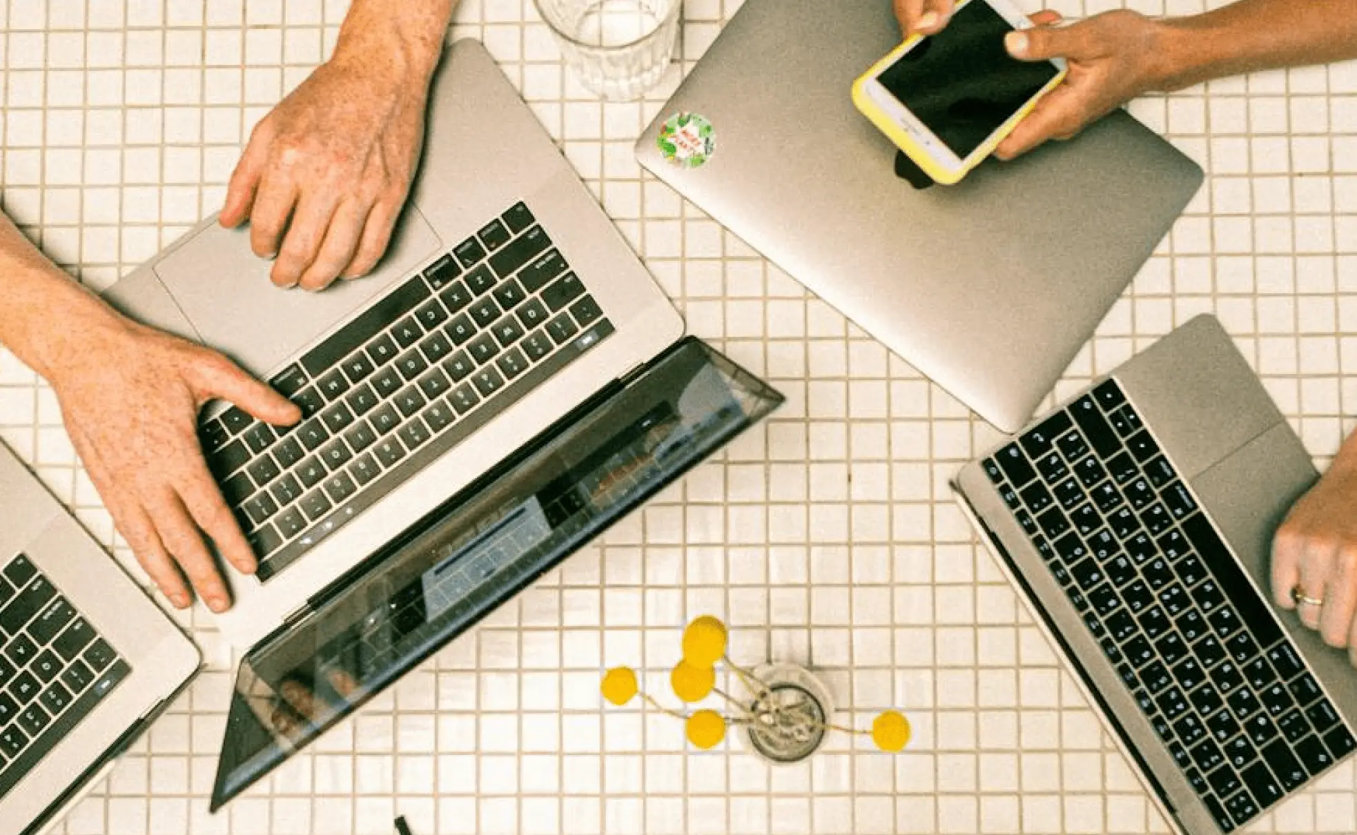 Top view of three people working at a table with laptops and a smartphone. A small vase with yellow flowers is on the table.
