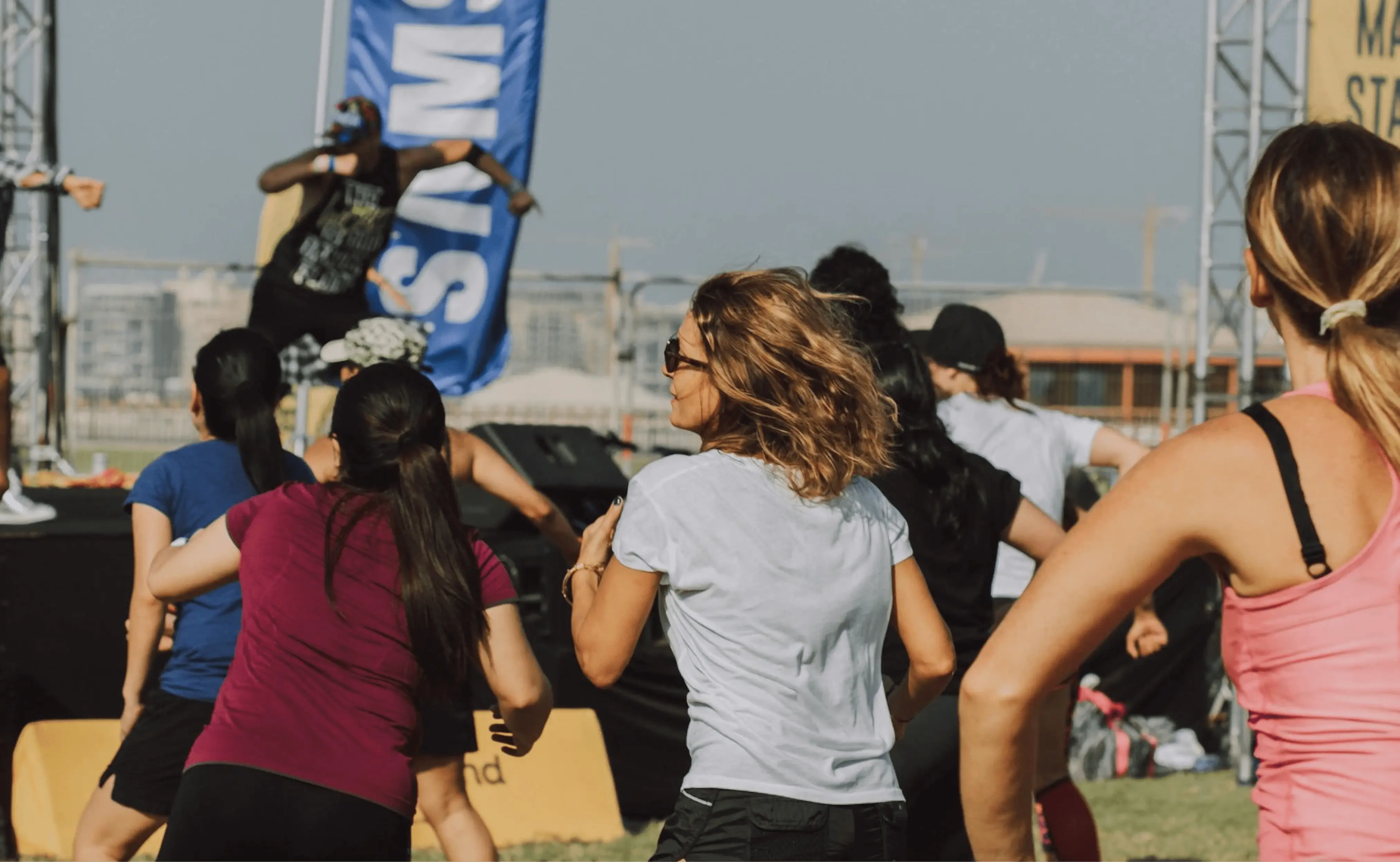 A group of people participating in an outdoor dance fitness class, actively moving to the music, led by an instructor on stage. The background includes cityscape and event banners.