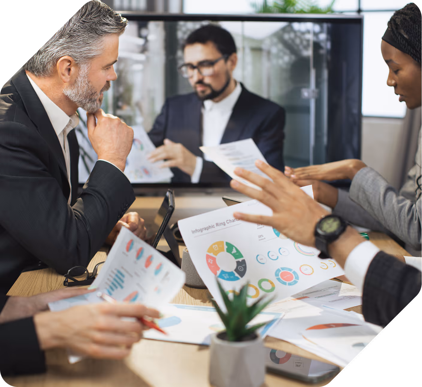 Business professionals in a meeting reviewing printed infographics and charts, with a man presenting on a screen in the background.