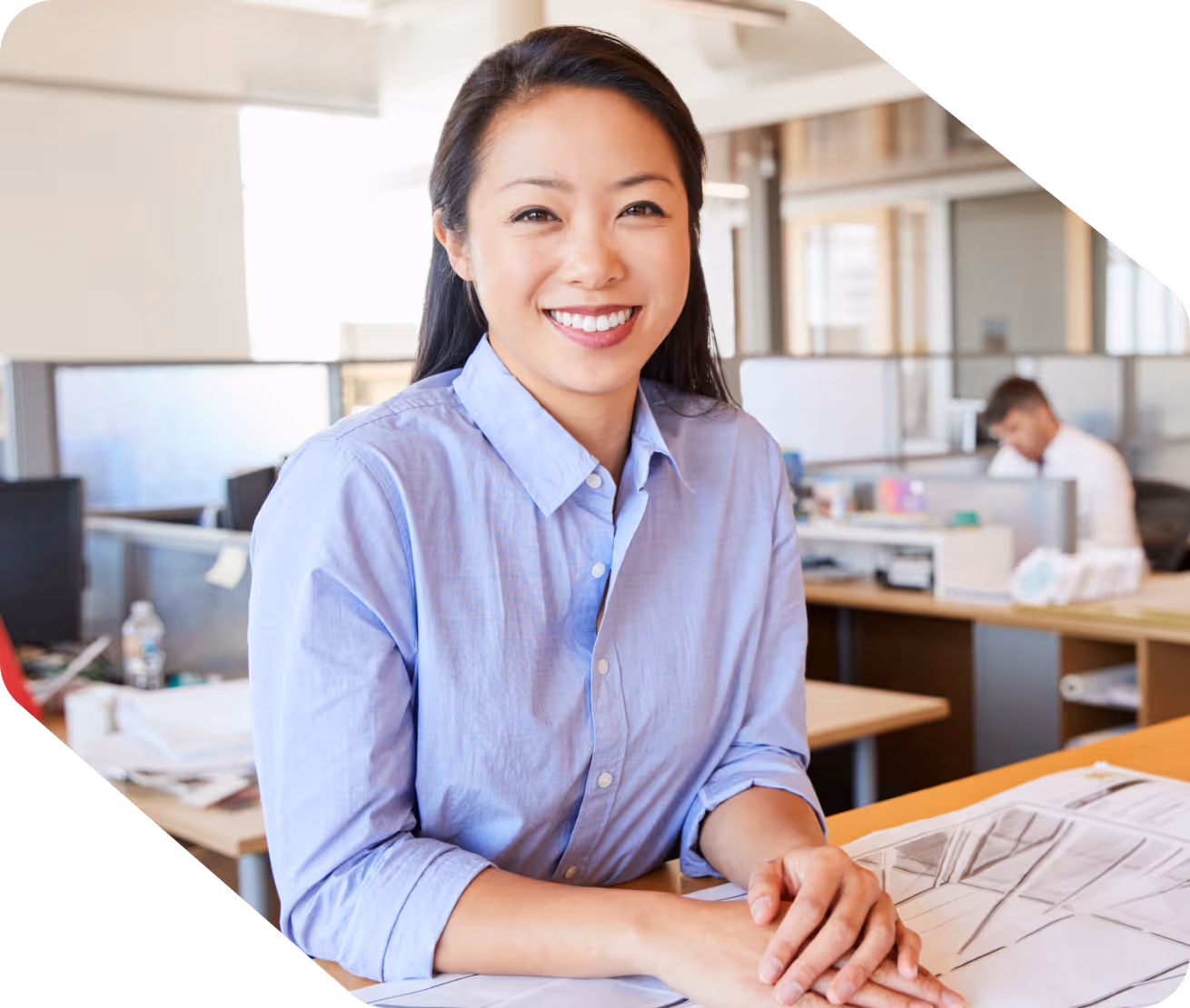 Smiling woman in a light blue shirt sitting at a desk with architectural plans in a bright office.