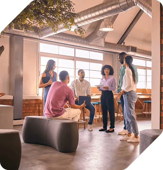 Group of six diverse young adults gathered in a modern office space having a discussion, some standing and some sitting on stools and a bench.