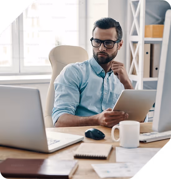 Man in blue shirt and glasses sitting at a desk holding a tablet, looking thoughtfully at a laptop.
