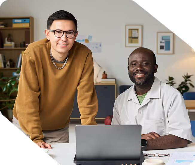 Two men smiling at a laptop in a modern office space with bookshelves and plants in the background.